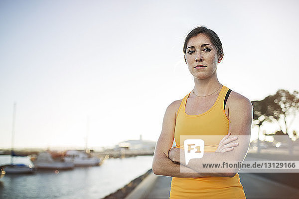 Porträt einer selbstbewussten Sportlerin  die mit verschränkten Armen am Hafen vor klarem Himmel steht
