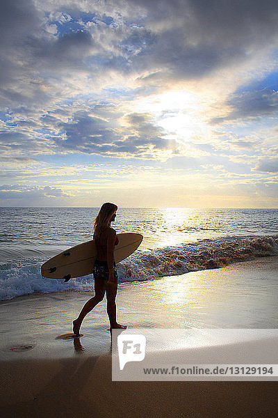 Frau mit Surfbrett geht am Meeresufer vor bewölktem Himmel bei Sonnenuntergang