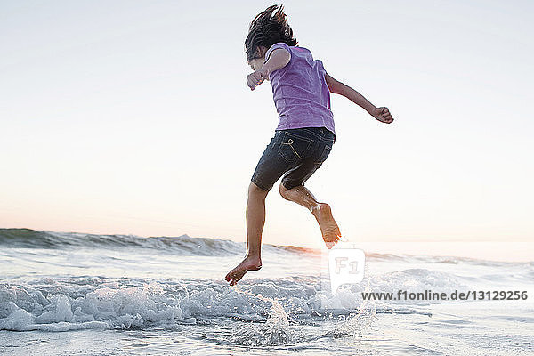 Tiefwinkelaufnahme eines Mädchens  das am Strand bei Sonnenuntergang auf Wellen gegen klaren Himmel springt