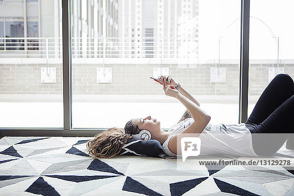 Side view of young woman using smart phone while lying on carpet by window at home