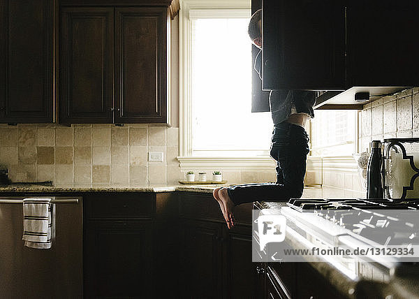 Boy searching something in cabinet while kneeling on kitchen counter at home