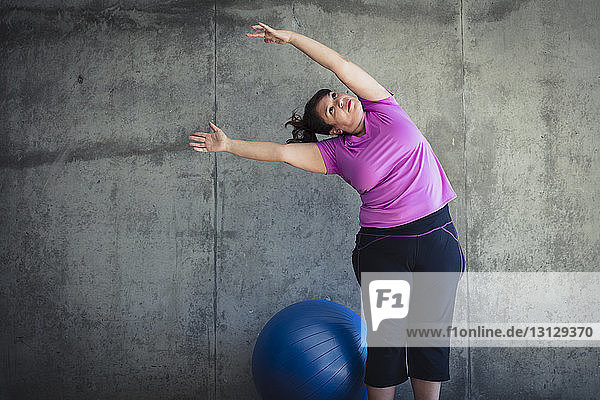 Frau mit erhobenen Armen übt Yoga  während sie beim Fitnessball an der Wand im Studio steht