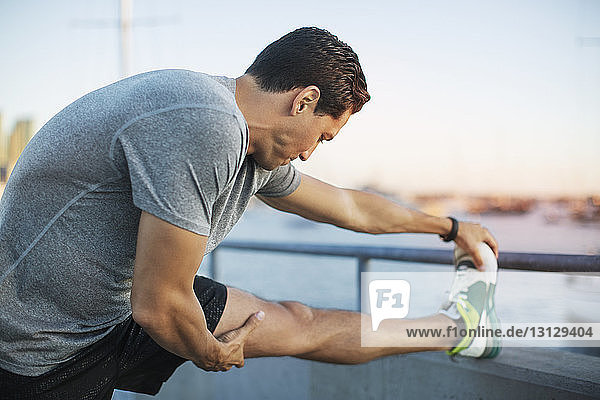 Side view of determined athlete stretching leg on pier by harbor