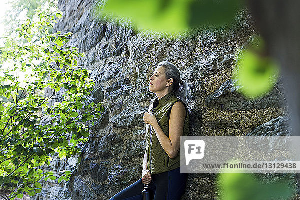 Woman closing zipper of jacket while leaning on stone wall