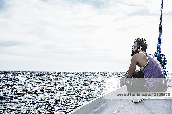 Seitenansicht eines Mannes  der auf das Meer blickt  während er auf einem Segelboot vor bewölktem Himmel sitzt