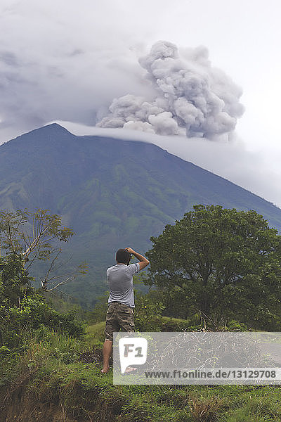 Rear view of man photographing smoke erupting volcanic mountain