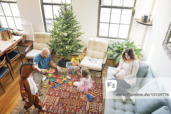 Hochwinkelaufnahme eines Vaters  der mit Kindern spielt  während eine schwangere Frau auf dem Sofa sitzt
