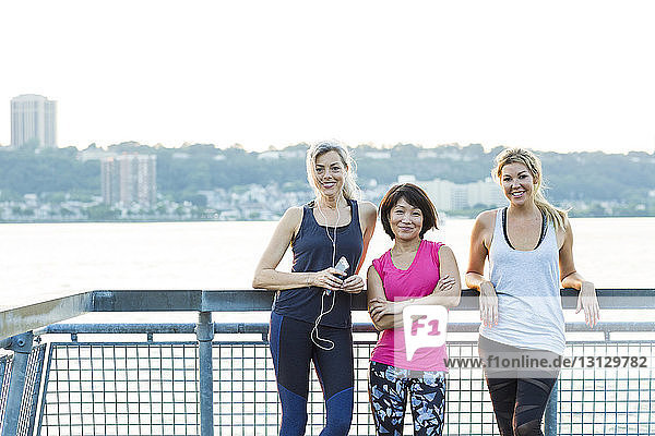 Portrait of confident female friends in sports clothing while standing on bridge against clear sky
