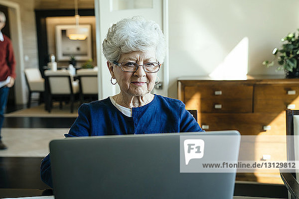 Ältere Frau benutzt zu Hause einen Laptop-Computer mit einem älteren Mann im Hintergrund