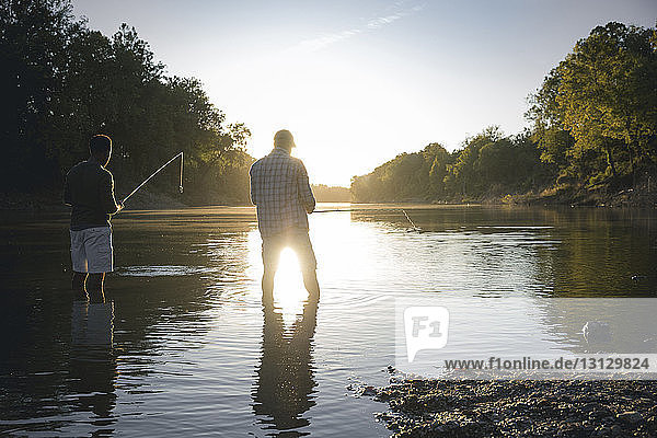 Rear view of male friends fishing in lake against sky during sunset