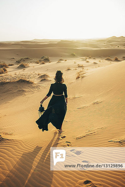 Rückansicht einer Frau  die während eines Spaziergangs in der Sahara bei Sonnenuntergang vor klarem Himmel die Laterne hält