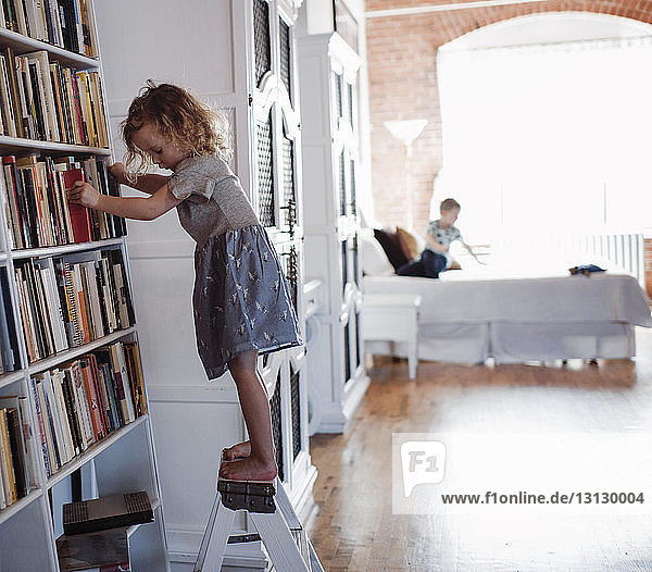 Side view of girl standing by bookshelves on ladder with brother in background at home