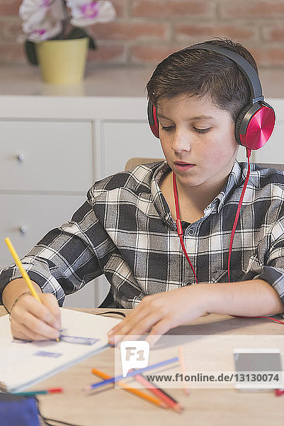 Junge hört Musik  während er zu Hause am Tisch mit Buntstiften ein Buch ausmalt