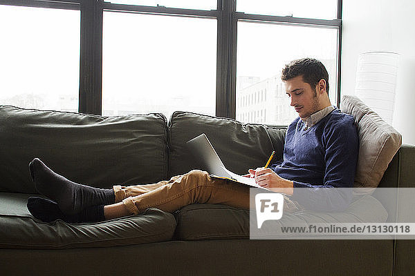 Side view of man using laptop and writing while sitting by windows