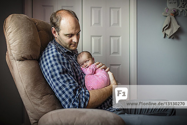 Vater trägt neugeborene Tochter  während er zu Hause auf einem Sessel sitzt