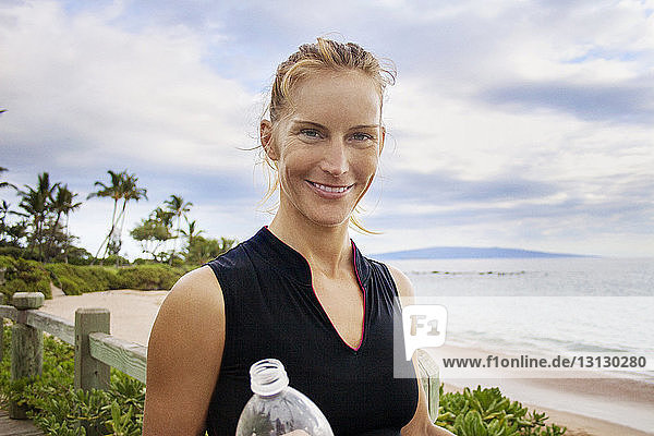 Portrait of smiling woman with bottle standing by beach against cloudy sky