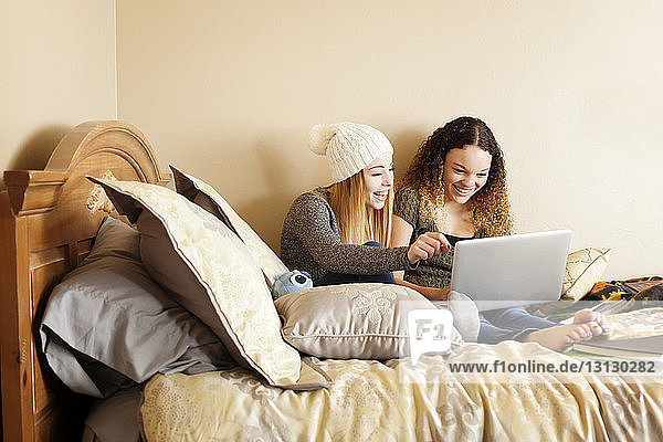 Happy friends using laptop computer while sitting on bed at home