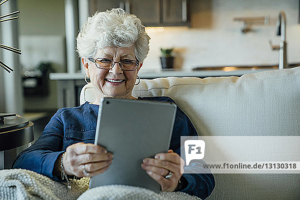 Ältere Frau lächelt  während sie einen Tablet-Computer auf dem Sofa zu Hause benutzt
