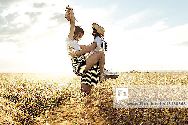 Happy woman carrying twin sister while standing on grassy field against sky