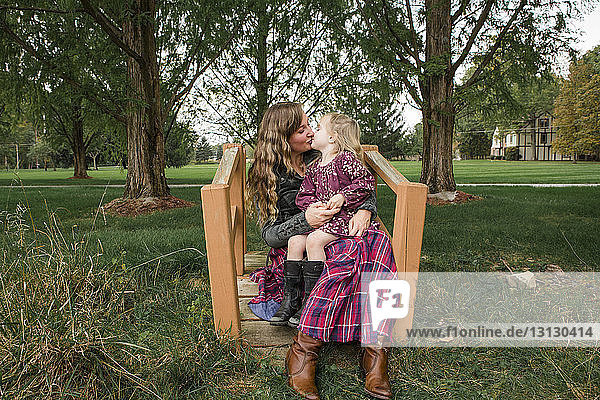 Tochter küsst Mutter  während sie im Park auf ihrem Schoß sitzt