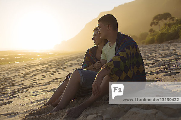 Junges Paar in eine Decke gewickelt  während es bei Sonnenuntergang auf den Strand schaut