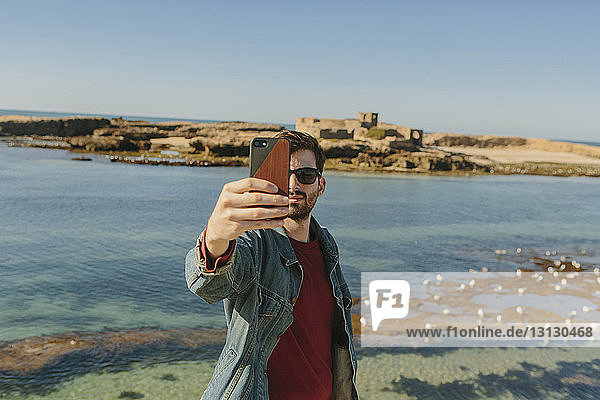 Handsome man taking selfie while standing at beach against clear sky