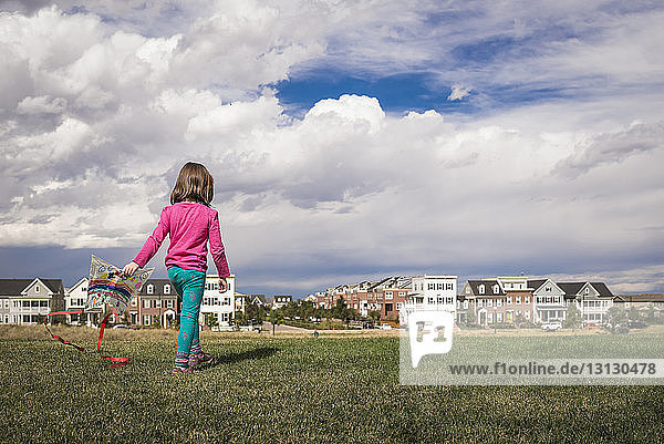 Rear view of girl with kite walking on grassy field against cityscape and cloudy sky
