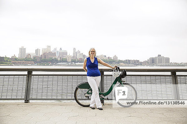 Porträt einer glücklichen  reifen Frau  die mit einem Fahrrad auf einer Promenade in der Stadt vor klarem Himmel steht