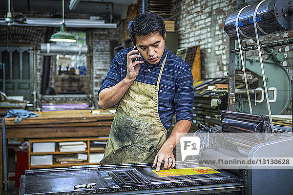 Mann benutzt Mobiltelefon  während er in der Werkstatt an der Druckmaschine steht