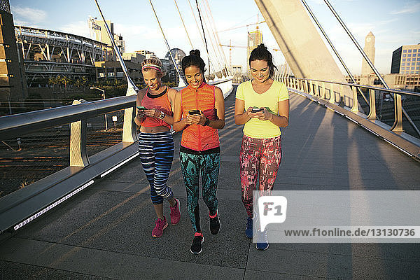 Smiling friends using phone while walking on bridge in city