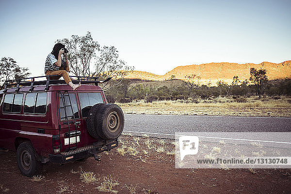 Woman photographing while sitting on car roof at desert