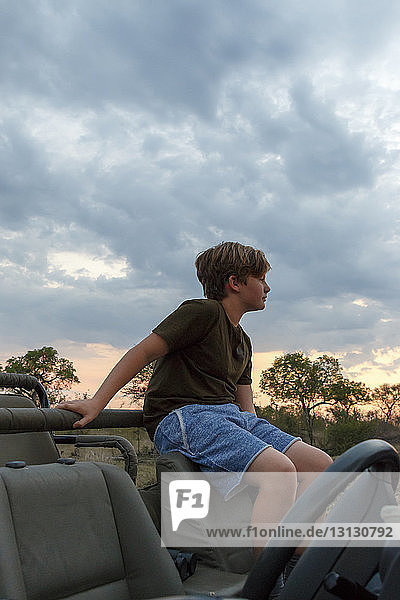 Thoughtful boy sitting on off-road vehicle against cloudy sky during sunset