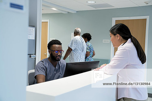 Arzt im Gespräch mit Krankenpfleger am Krankenhausempfang  während Kollege mit Patient im Hintergrund geht