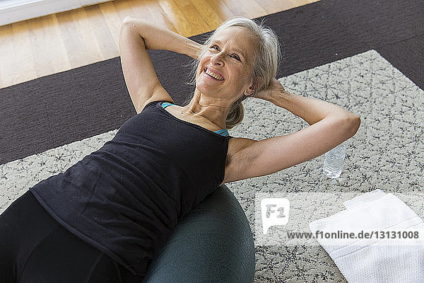 Lächelnde ältere Frau mit Händen hinter dem Kopf auf dem Fitnessball liegend während sie zu Hause Sport treibt