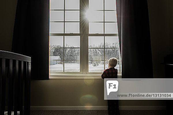 Rear view of boy looking through window while standing at home during winter