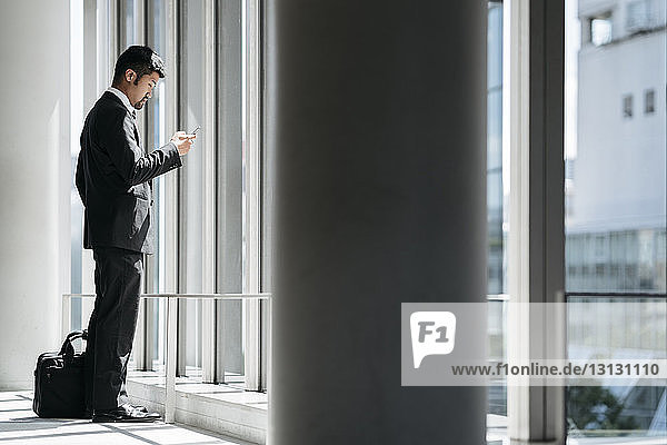 Seitenansicht eines Geschäftsmannes  der ein Smartphone benutzt  während er im Büro am Fenster steht