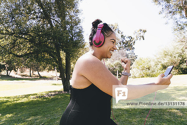 Seitenansicht einer glücklichen Frau  die ein Smartphone betrachtet  während sie im Park Musik hört