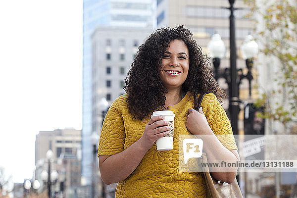 Smiling woman holding disposable cup while standing at sidewalk
