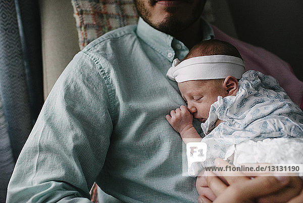 Midsection of father carrying sleeping newborn daughter while sitting on chair at home