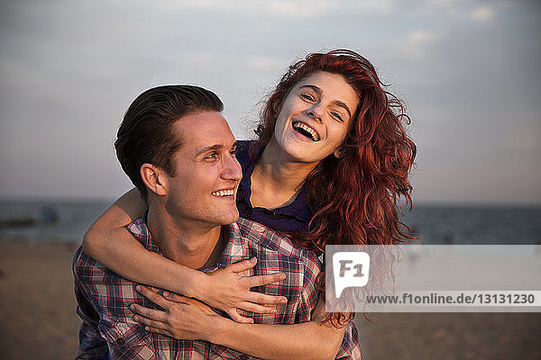 Glücklicher Mann nimmt Frau bei Sonnenuntergang am Strand huckepack