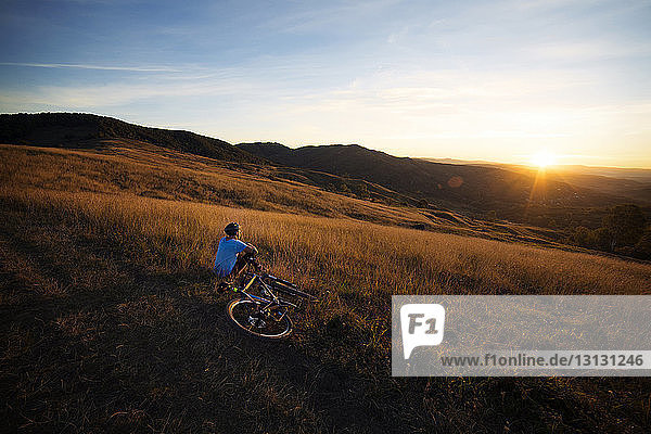Male athlete with bicycle sitting on grassy field against sky during sunset