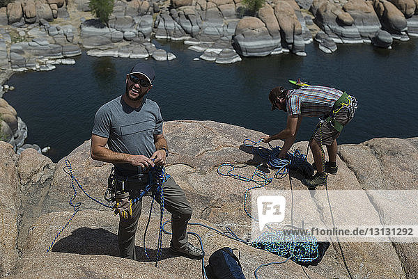 Hochwinkelaufnahme von Freunden  die bei Sonnenschein Kletterseile binden  während sie auf einer Felsformation über dem See stehen