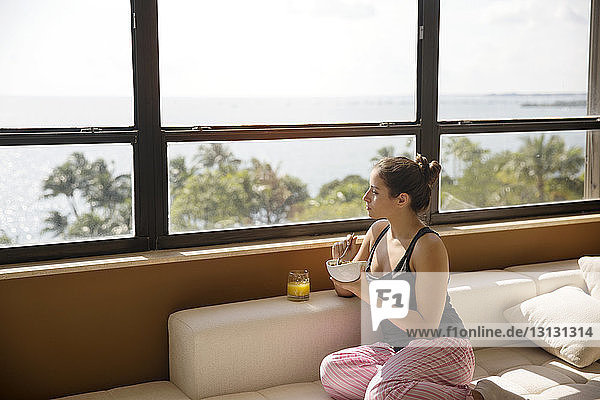 Frau schaut durchs Fenster  während sie zu Hause auf dem Sofa frühstückt