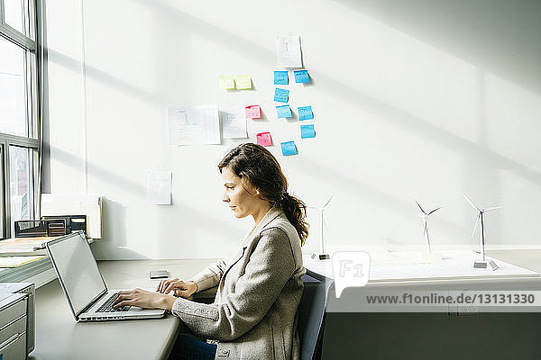 Seitenansicht einer Geschäftsfrau  die einen Laptop benutzt  während sie an auf dem Schreibtisch angeordneten Modellen von Windkraftanlagen arbeitet