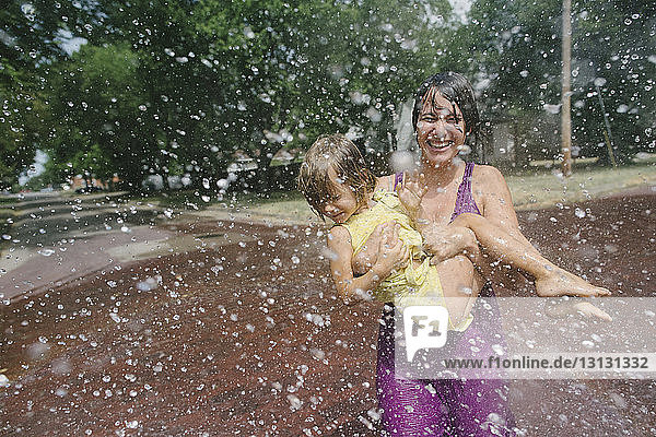 Verspielte Mutter  die ihre Tochter austrägt  während sie sich im Springbrunnen im Park vergnügt