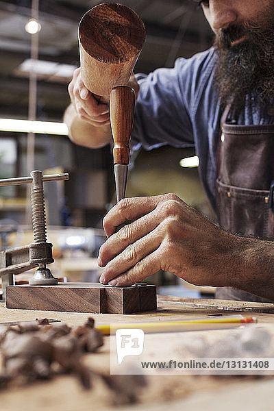 Beschnittenes Bild eines Handwerkers  der in einer Holzwerkstatt Werkzeuge in ein Brett hämmert