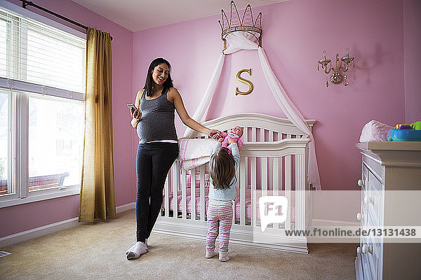 Lächelnde schwangere Frau benutzt Smartphone  während sie zu Hause ihre verspielte Tochter ansieht
