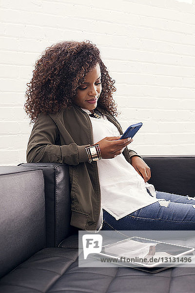 Smiling businesswoman using phone while sitting by digital tablet on sofa at office