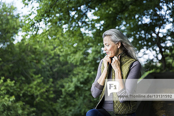 Thoughtful woman holding jacket while leaning on retaining wall against branches