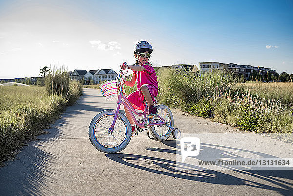 Girl riding bicycle with training wheels on road against sky during sunny day